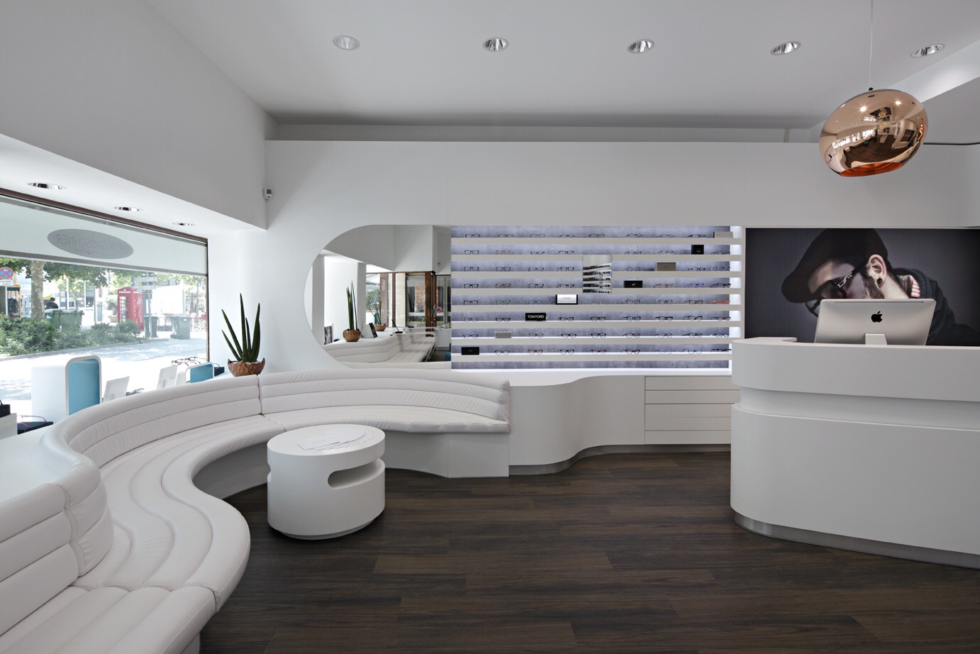 Helle, moderne Empfangshalle mit geschwungener weißer Sitzbank, rundem weißen Tisch, dunklem Holzboden und einem Empfangstresen mit Computer und Porträt an der Wand.