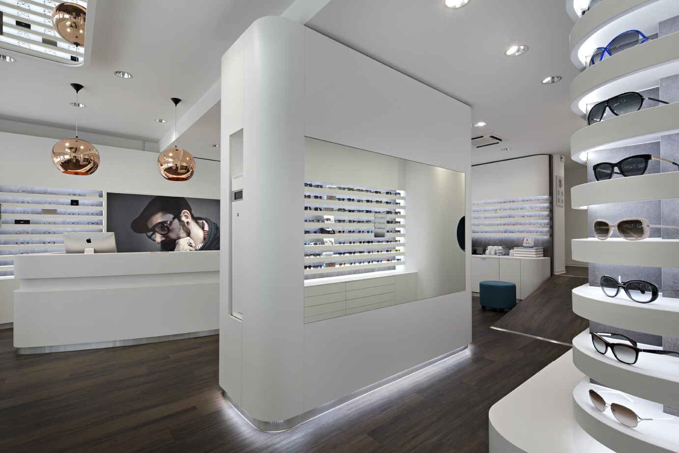 Optik Gröschner in Bruchsal Innenraum eines modernen Brillengeschäfts mit weißen Wänden, dunklem Holzboden und mehreren Brillenpräsentationen an Wänden und runden Ständern.