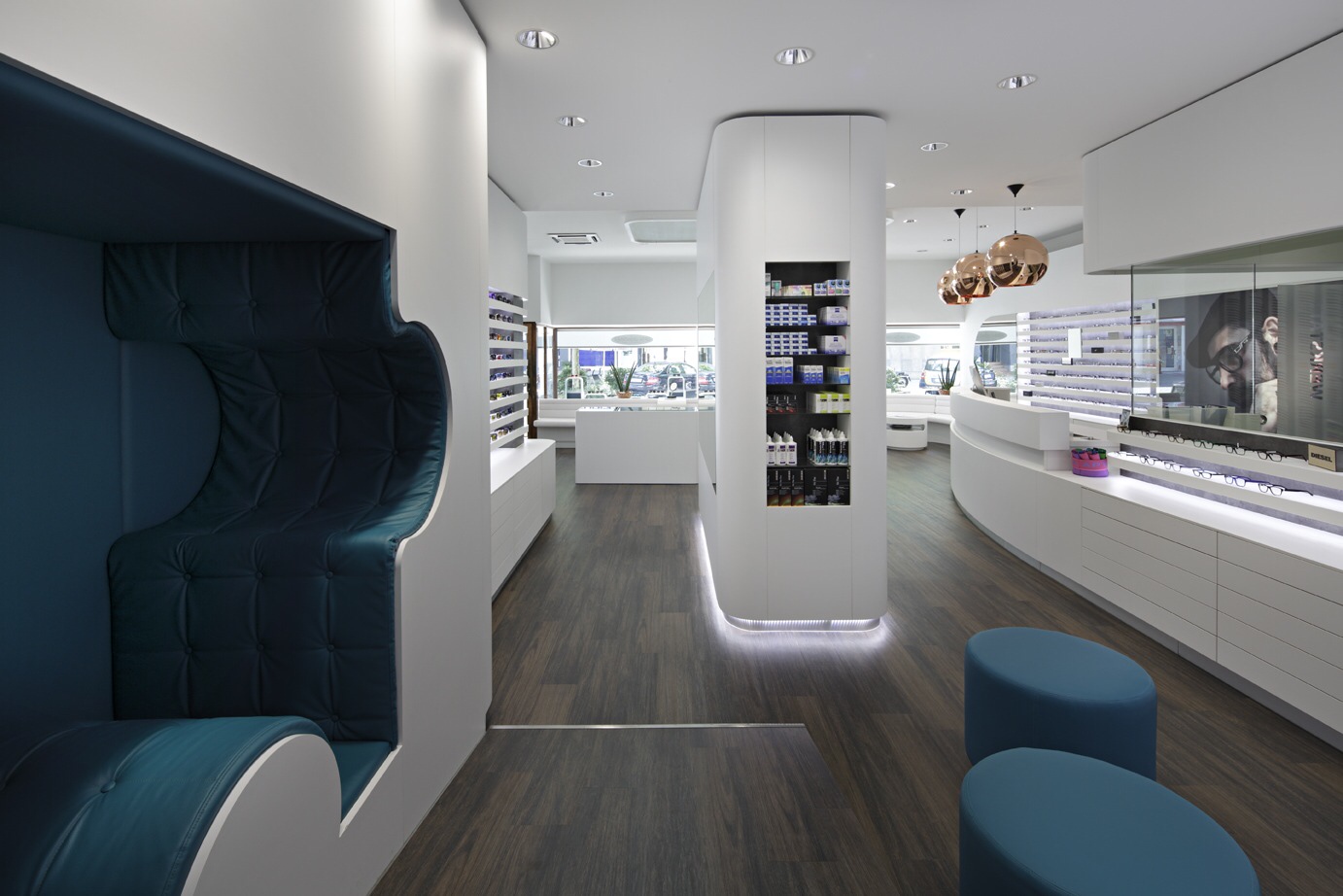 Optik Gröschner in Bruchsal Moderner Verkaufsraum mit geschwungenen weißen Theken, dunklem Holzboden, blauen Sitzhockern und einer beleuchteten Säule mit Regalen
