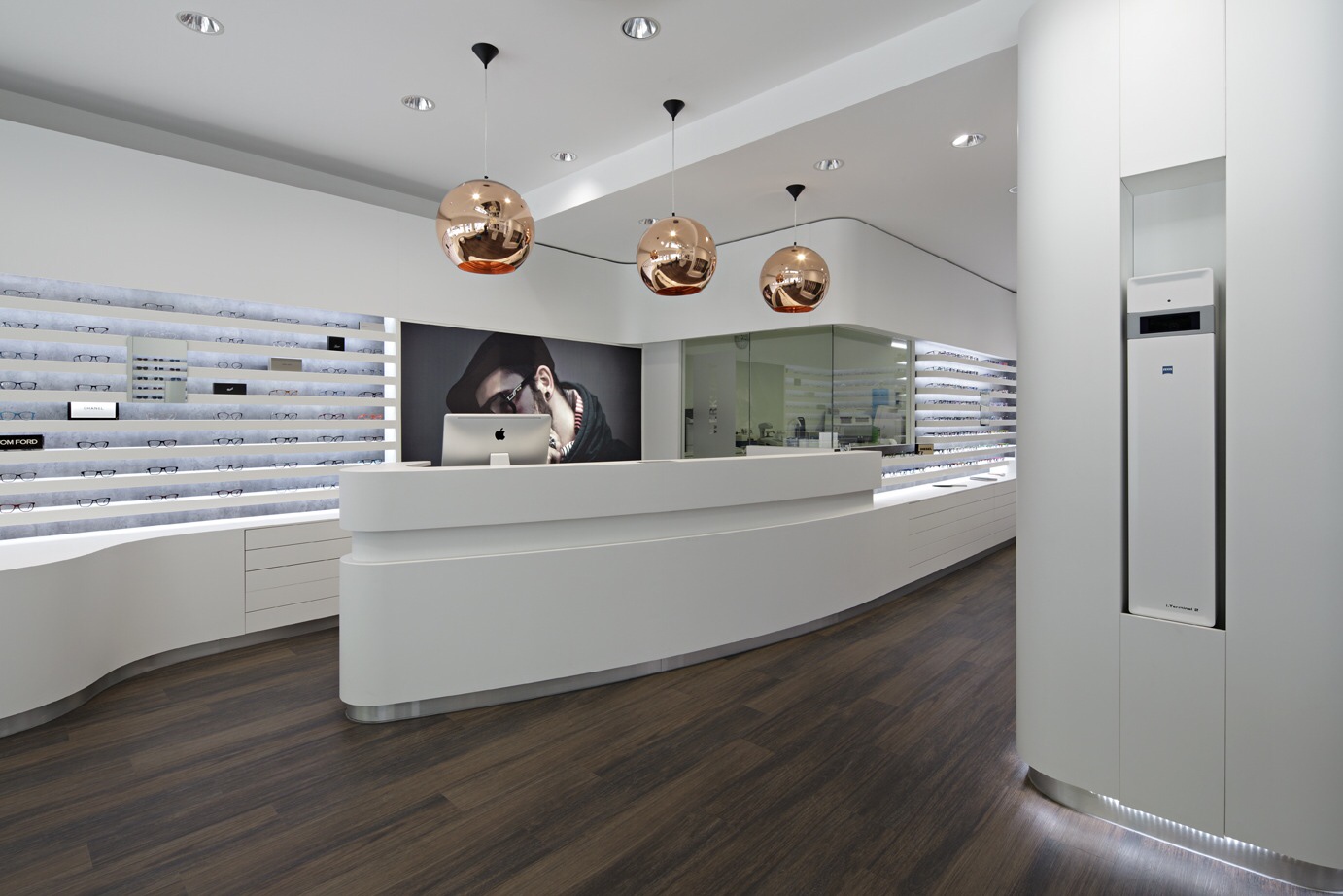 Optik Gröschner in Bruchsal Moderner Empfangsbereich mit geschwungener weißer Theke, drei kupferfarbenen Hängeleuchten und großem Porträt an der Wand.