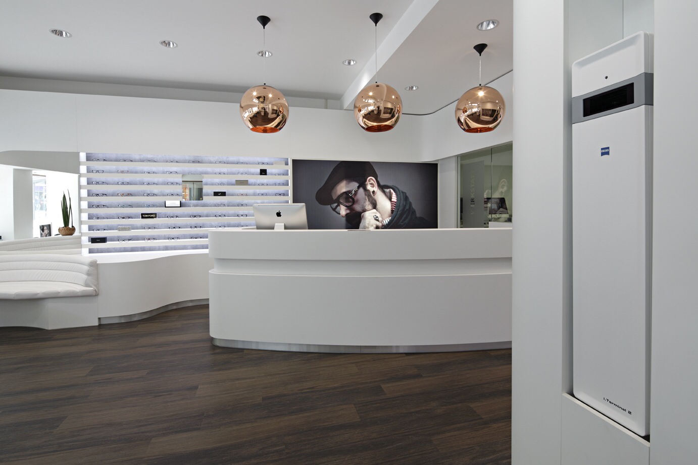 Optik Gröschner in Bruchsal Moderne Empfangstheke mit drei kupferfarbenen Pendelleuchten und großem Porträt eines Mannes mit Brille und Bart im Hintergrund