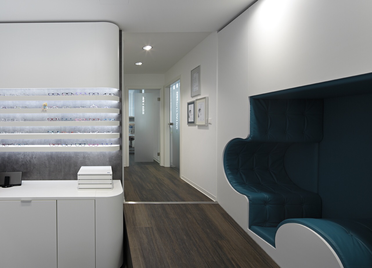 Optik Gröschner in Bruchsal Moderner Innenraum mit weißem Empfangstresen, dunklem Holzboden, weiß gestrichenen Wänden, beleuchtetem Regal und einer blauen Sitznische mit abgerundeter Form.