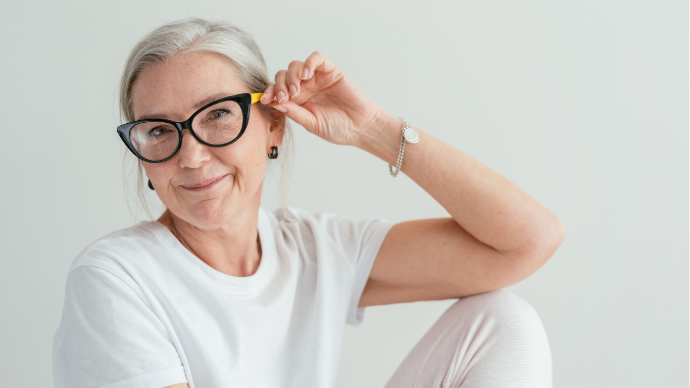 Gleitsichtbrille Ältere Frau mit grauen Haaren, die eine schwarze Gleitsichtbrille trägt und lächelnd eine Brillenbügelspitze hält.