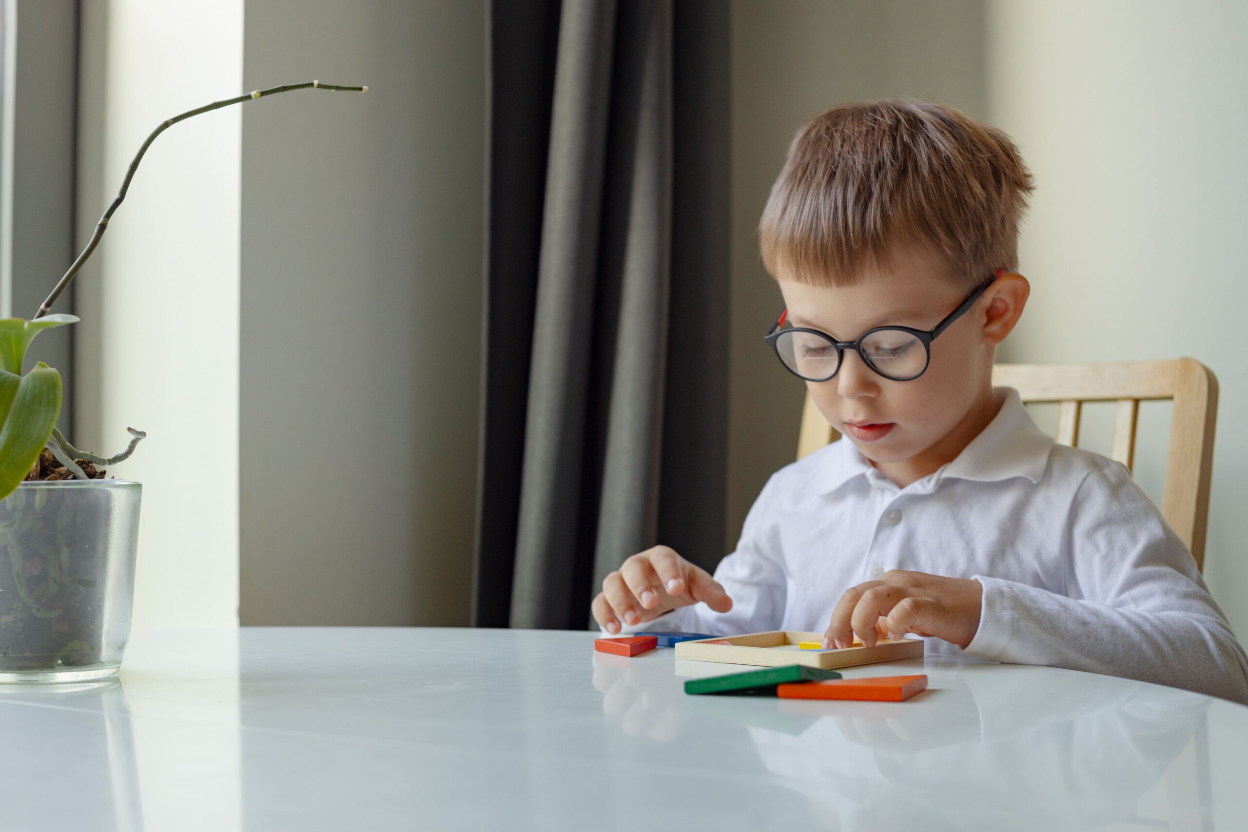 Kinderbrille Junge mit Brille sitzt an einem weißen Tisch und spielt mit bunten Holzbausteinen