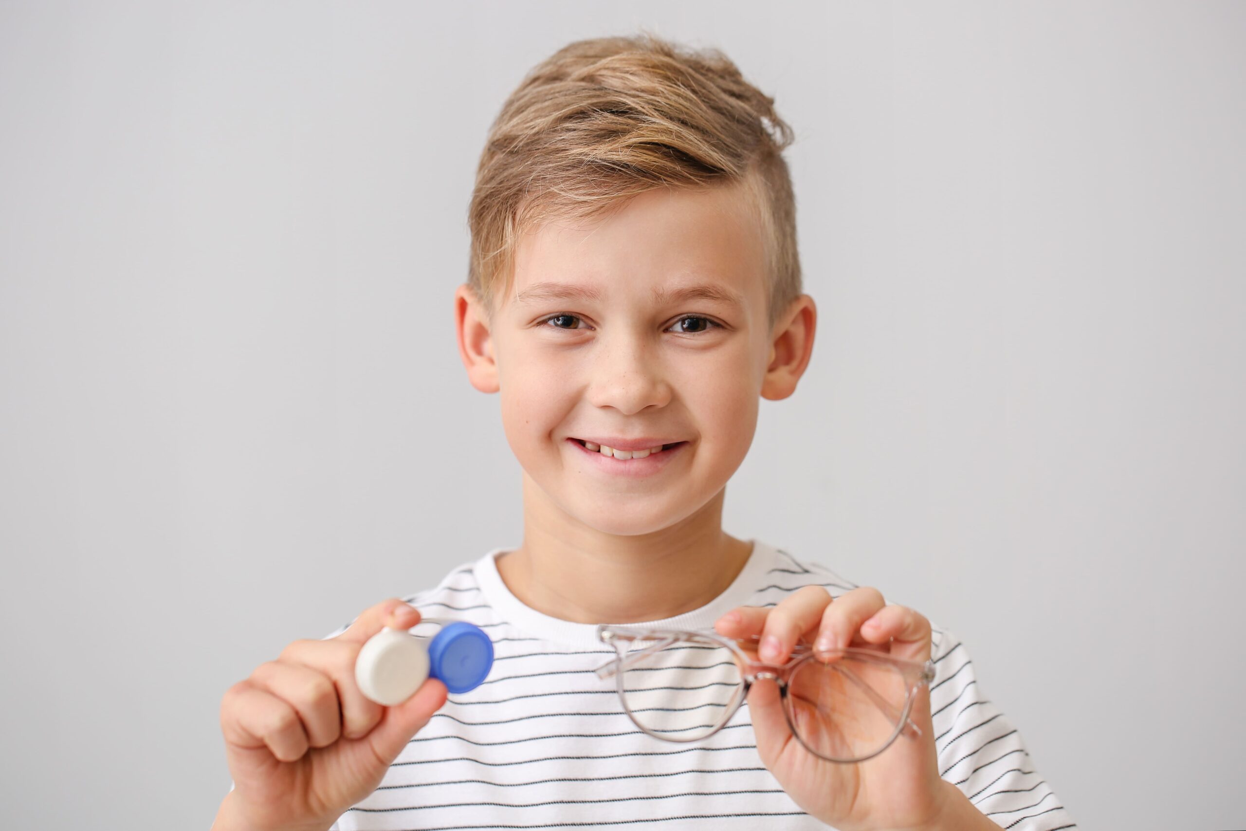 Kurzsichtigkeit aktiv kontrollieren Lächelnder Junge mit gestreiftem T-Shirt hält eine Brille in der einen und ein Kontaktlinsenbehälter in der anderen Hand.