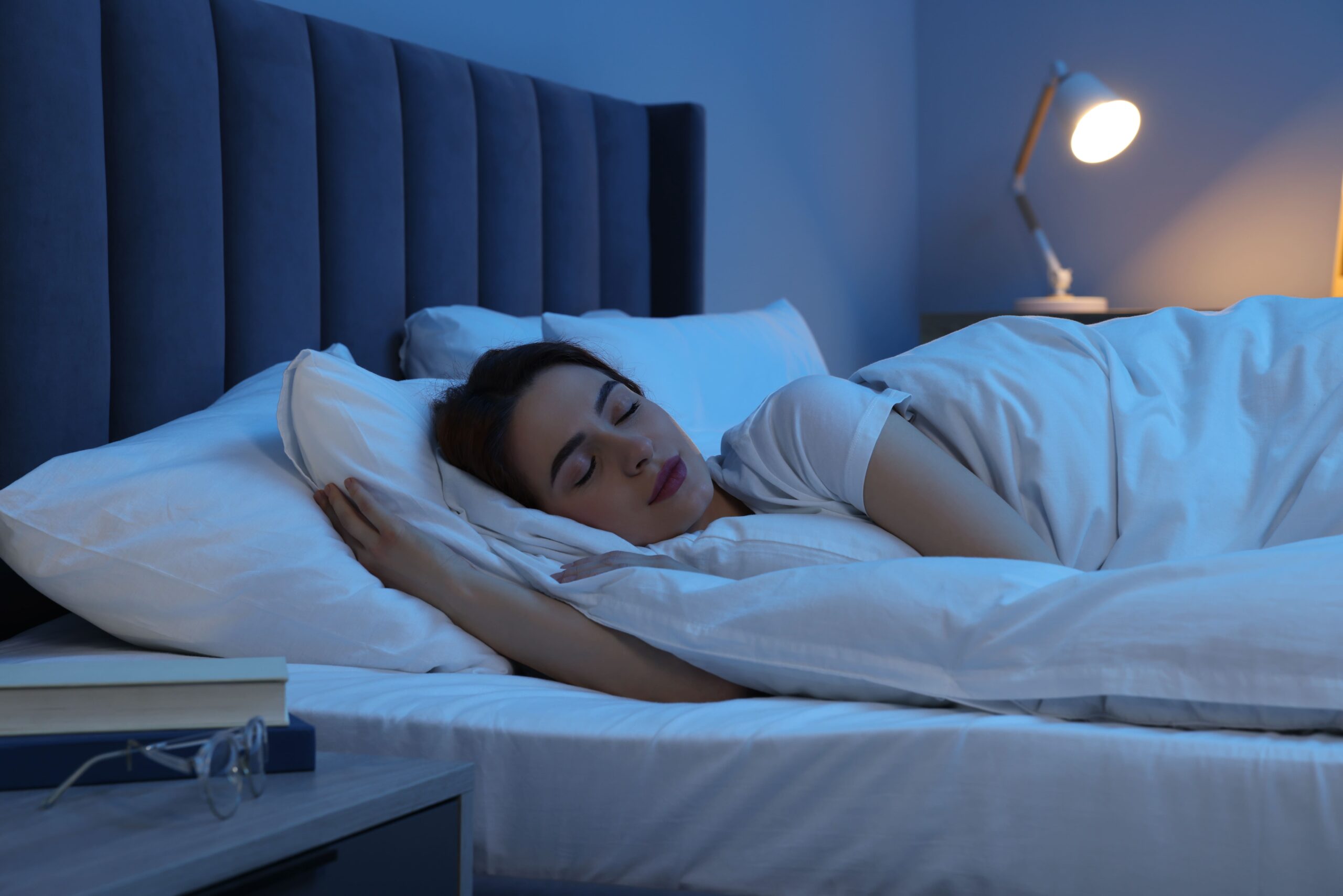 Ortho-K-Kontaktlinsen Frau liegt schlafend auf der Seite in einem Bett mit gepolstertem Kopfteil und weißer Bettwäsche, neben dem Bett steht ein Nachttisch mit Büchern und einer Brille, im Hintergrund eine eingeschaltete Leselampe.