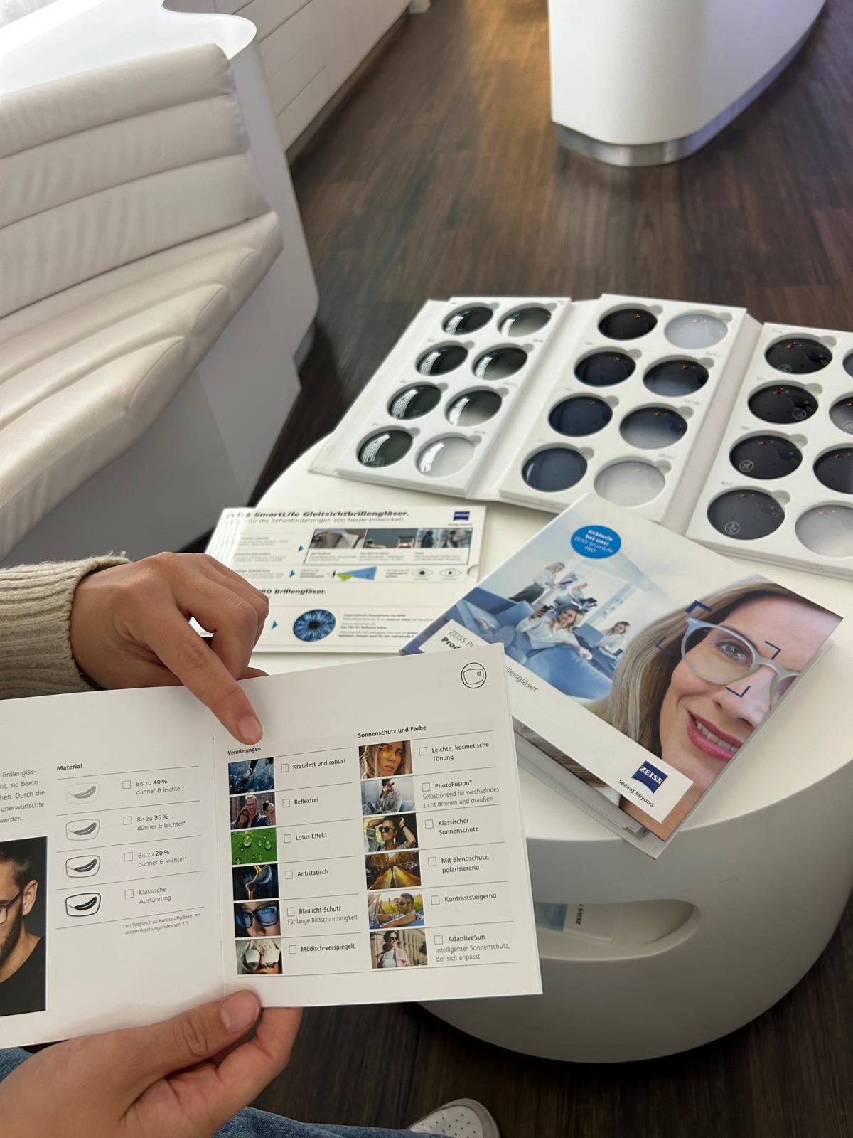 Unsere Services Person hält eine Broschüre mit Bildern und Texten, auf einem Tisch liegen mehrere Tabletts mit Kontaktlinsen und eine weitere Broschüre mit einem Porträt einer Frau.