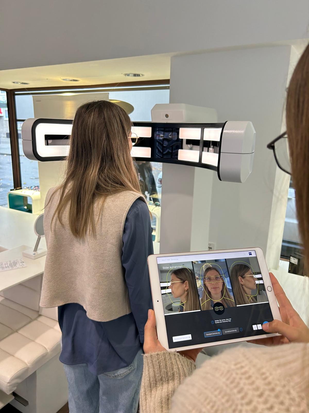 Frau steht vor einem Gesichtsscanner, eine andere Person hält ein Tablet mit drei Porträtfotos der Frau.