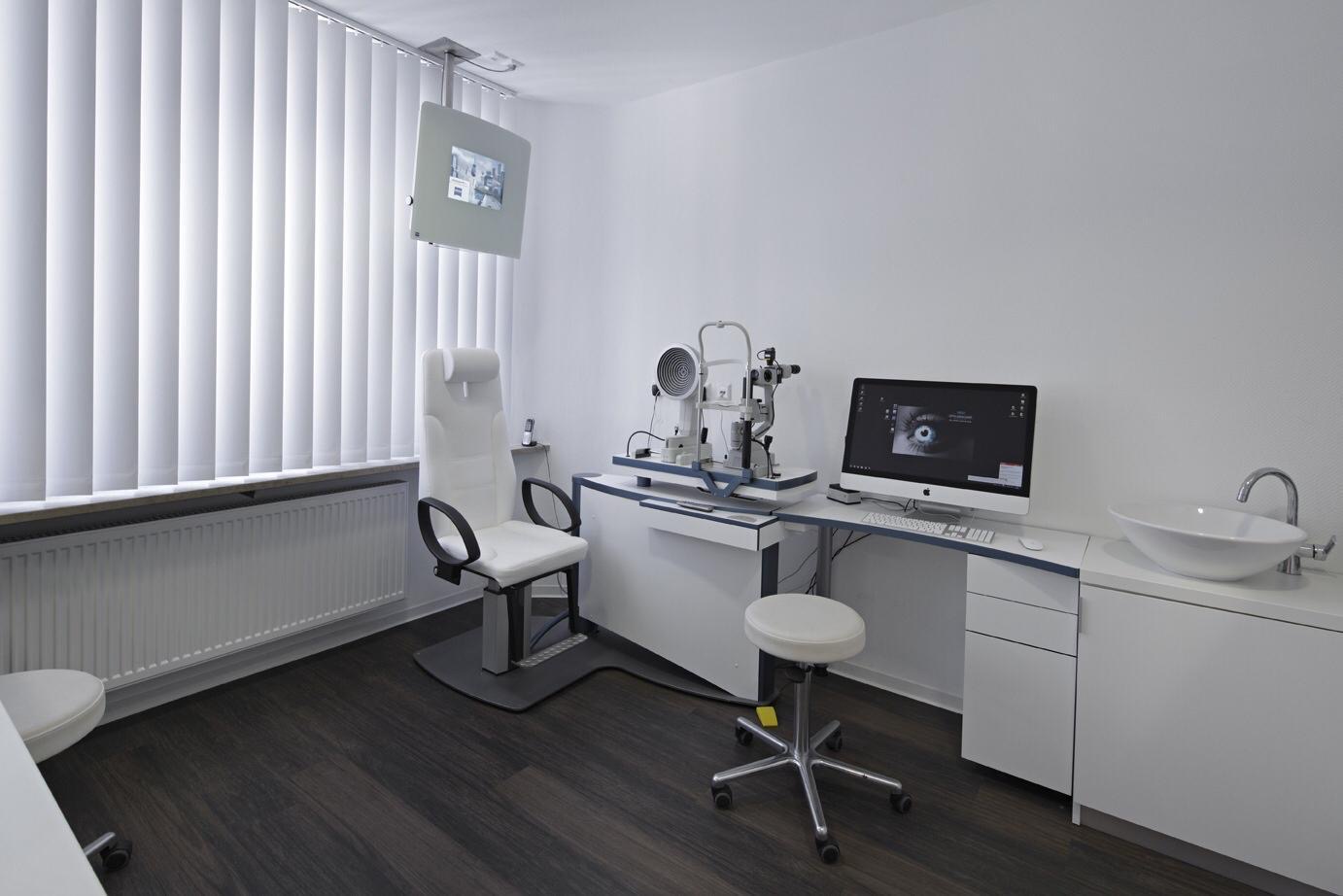 Warum ist ein Augenscreening sinnvoll? Behandlungszimmer mit Augenuntersuchungsgerät, Stuhl, Hocker, Computerbildschirm und Waschbecken.