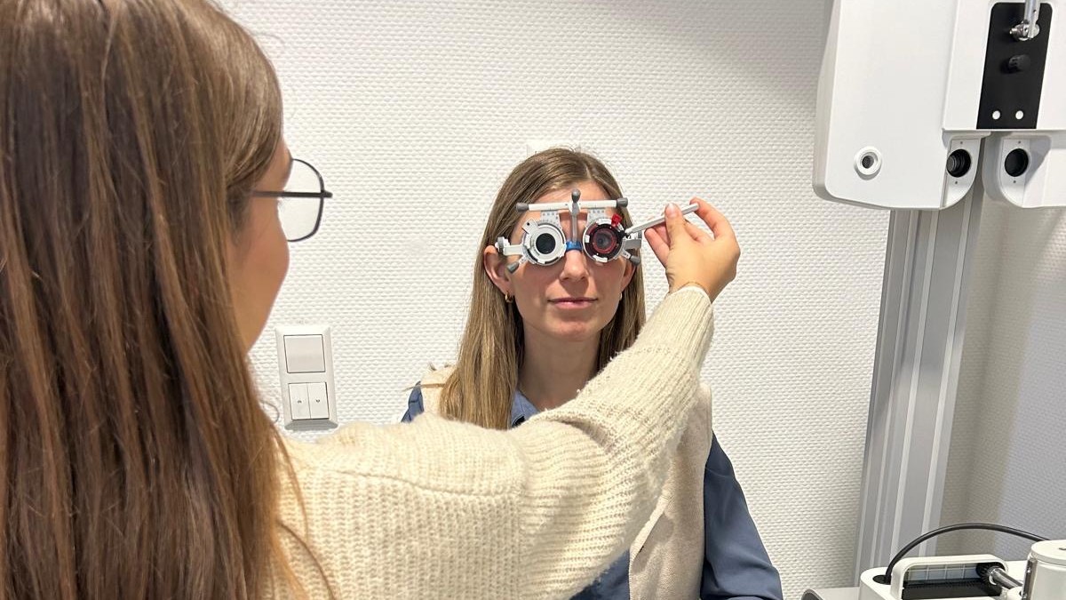 Frau mit Brille stellt bei einer anderen Frau eine Sehhilfe vor deren Augen ein, die zweite Frau trägt eine blaue Bluse und sitzt vor einer weißen Wand